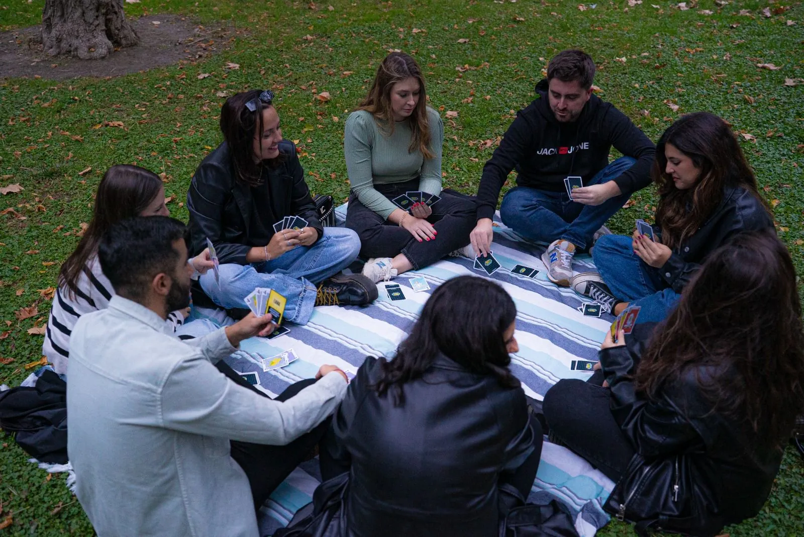 Ocho personas sentadas en círculo sobre una manta en un parque, jugando una partida de Rey Mono con las cartas repartidas por el suelo